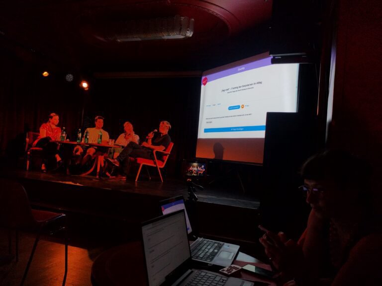 The photo shows the use of the interaktive event module at a panel discussion. On the left there the peope who spaek at the panel with the questions from the modul in the projected background.